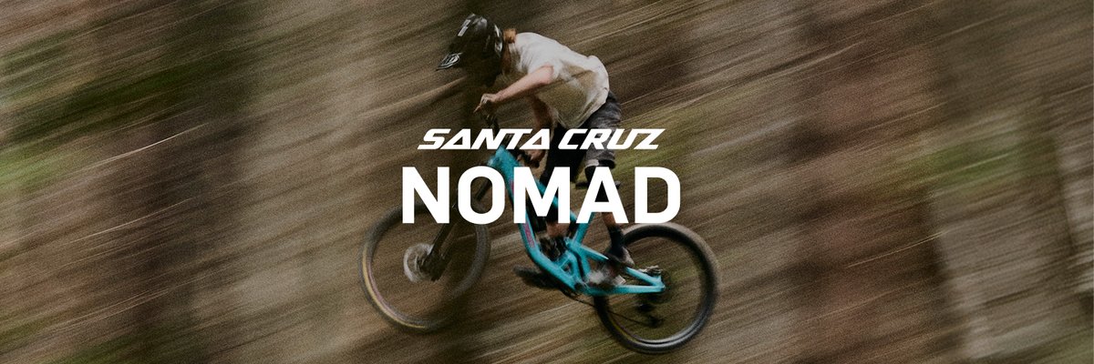 A rider on a teal Santa Cruz Nomad mountain bike, captured mid-descent through a wooded trail with heavy motion blur conveying speed.
