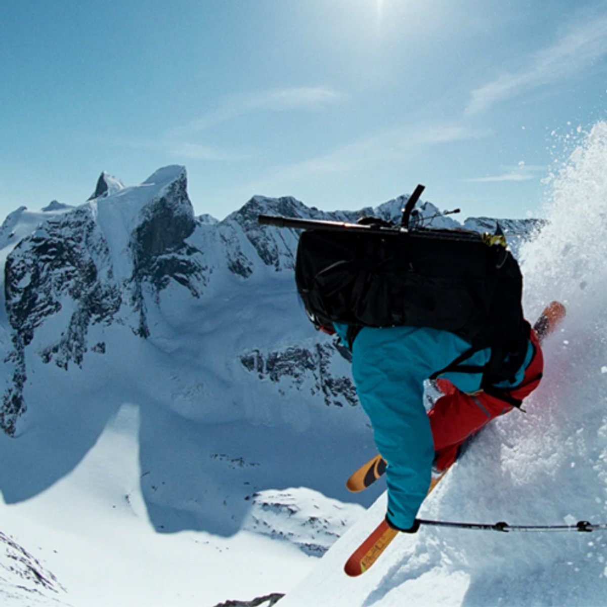 A skier in turquoise and red gear performs an aerial jump off a snowy mountain peak with dramatic alpine peaks visible in the background.
