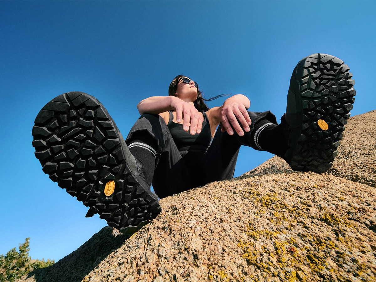 Underside view of two Bedrock Mountain Clogs facing camera, showing Vibram Megagrip lug pattern and yellow Vibram medallion, worn by person reclining on granite rock against blue sky