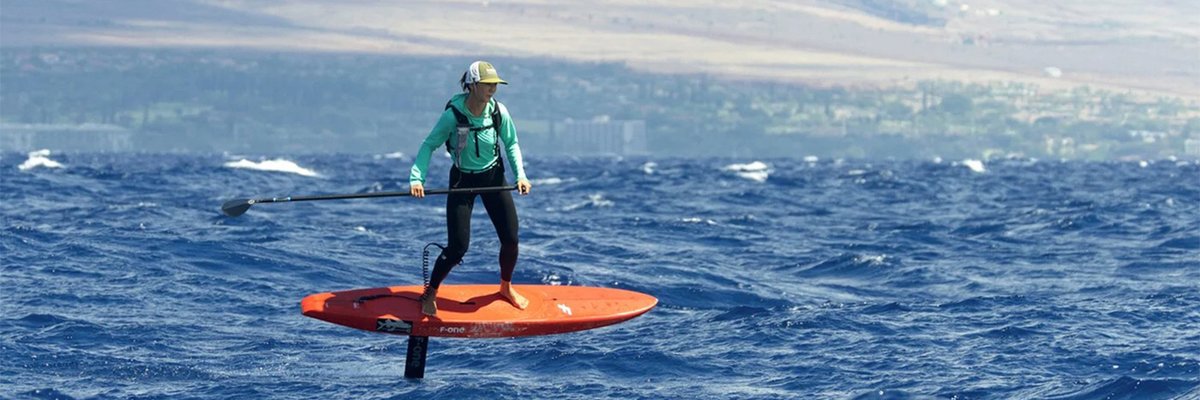A stand-up paddleboarder in a green wetsuit paddles an orange board on blue ocean waters with a volcanic mountain landscape in the background.