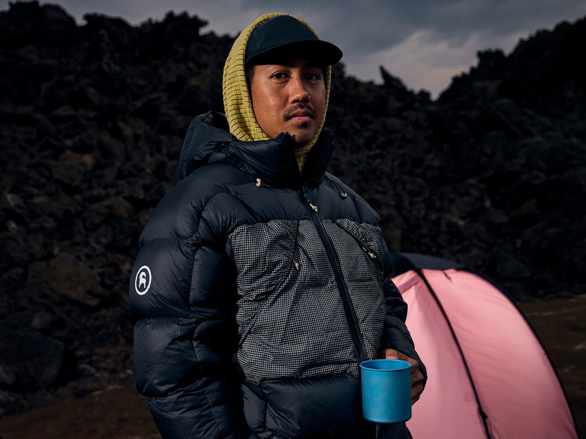 A person in a navy puffer jacket and yellow hood holds a blue camp mug beside a pink tent, dark volcanic rock looming behind them.