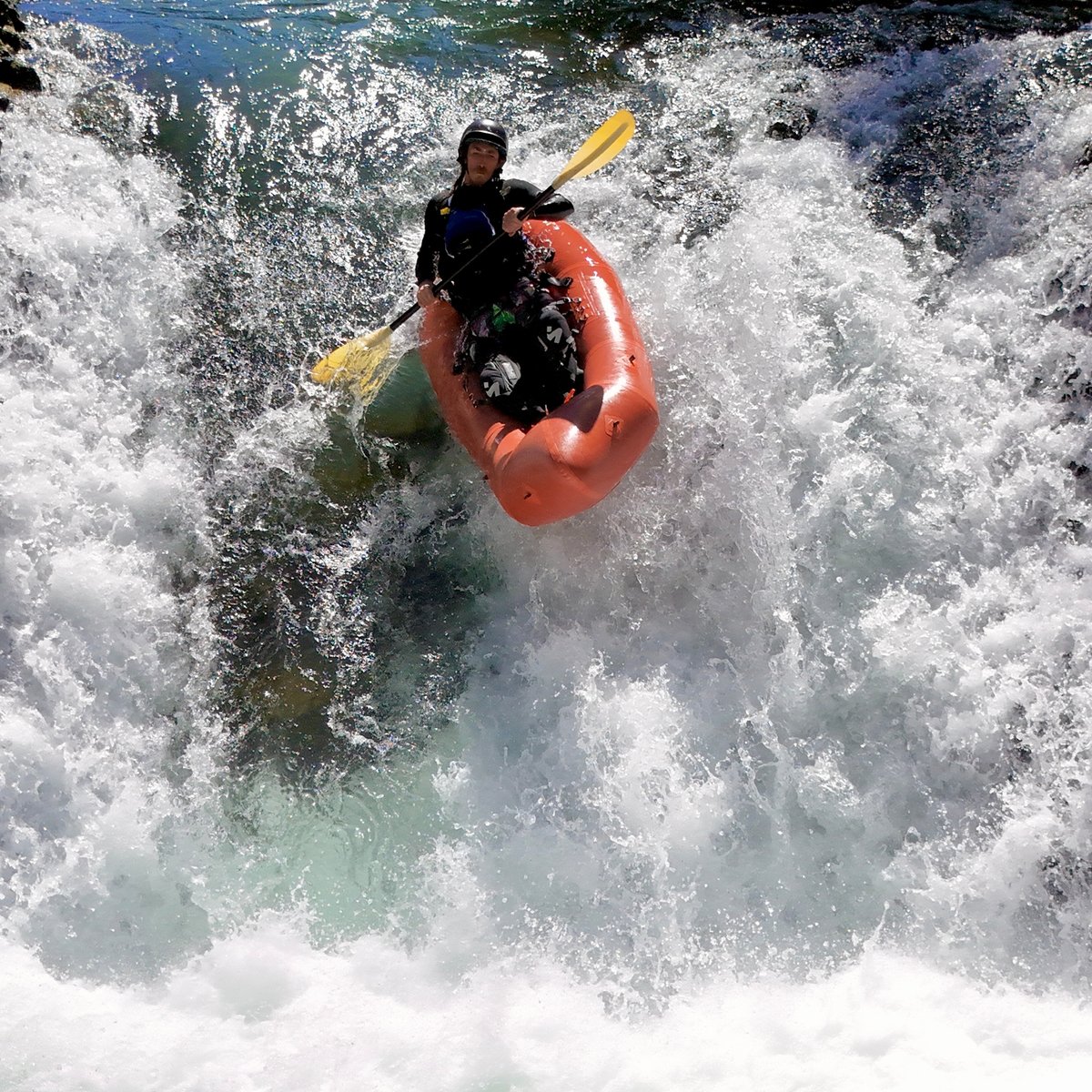 A person in an orange inflatable kayak paddles down a whitewater waterfall, creating a large splash.