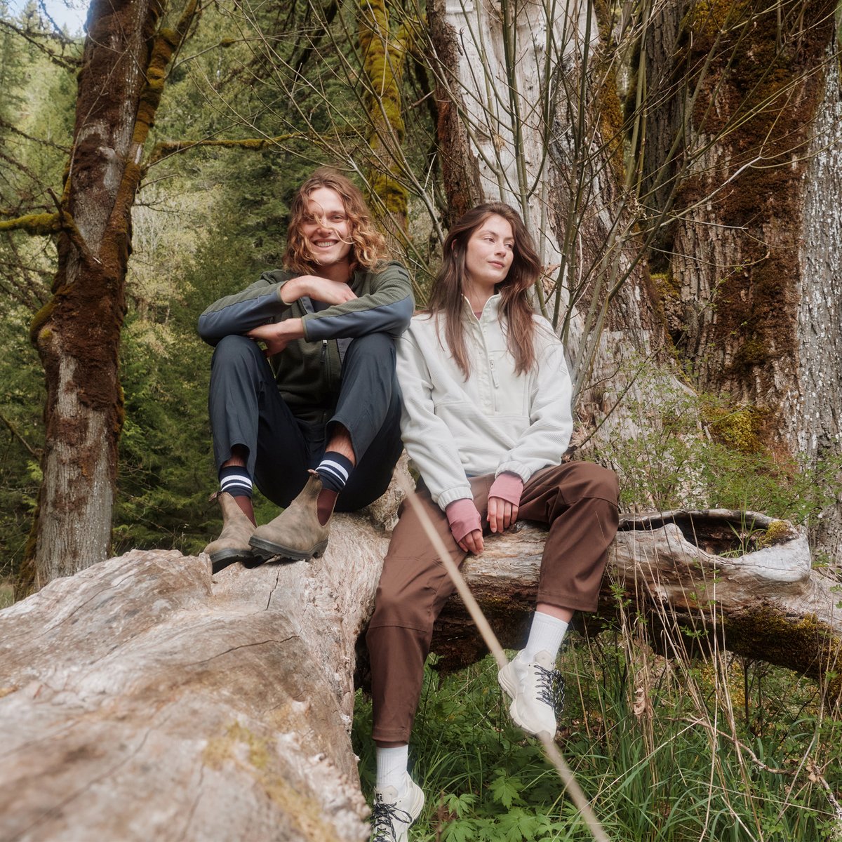 A man and a woman sit on a fallen tree in a forest, each wearing prAna Stretch Zion pants.