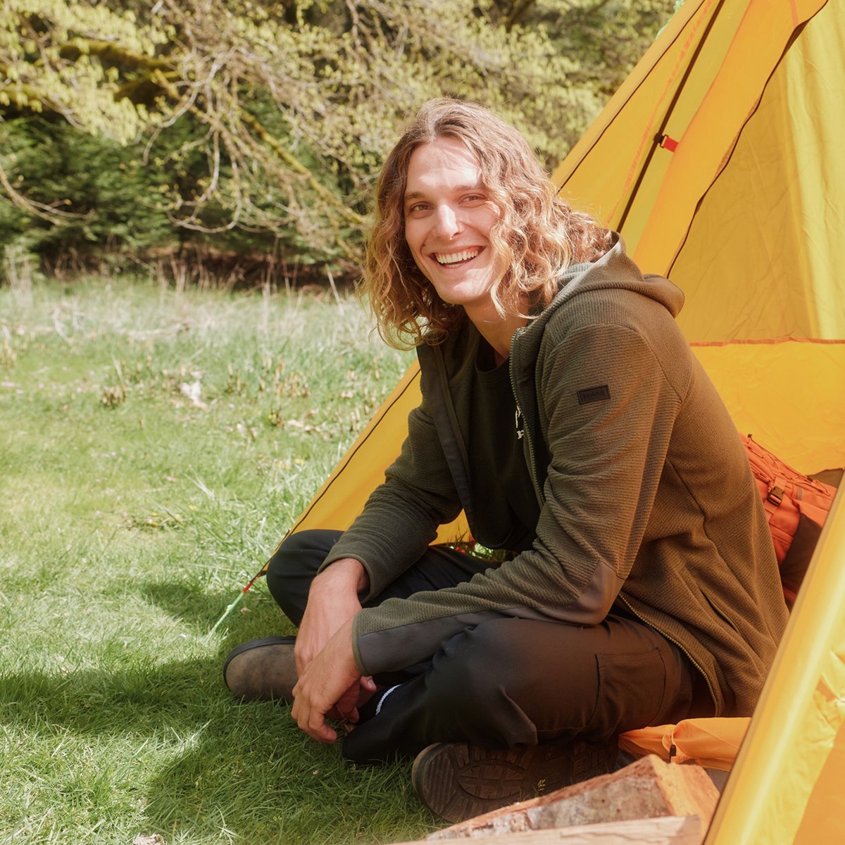 A man sits in the open door of a tent, smiling in the sunshine as he wears the prAna Zion Stretch Pant.