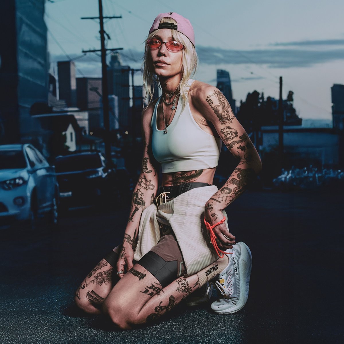 A person with tattooed arms and legs kneels on a city street at dusk, wearing a white sports bra, 2-layer shorts, a pink cap and sunglasses, a sweatshirt tied around the waist, and white Salomon sneakers.