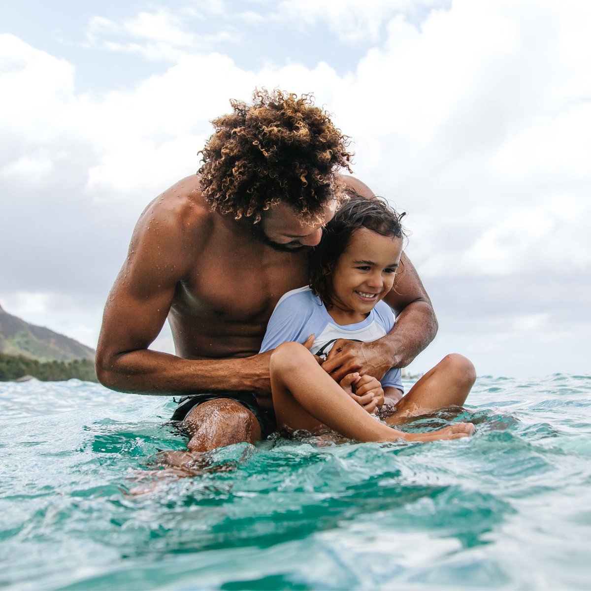 Father and young child playing together in clear turquoise ocean water on a tropical beach with mountains in the background during a sunny day.