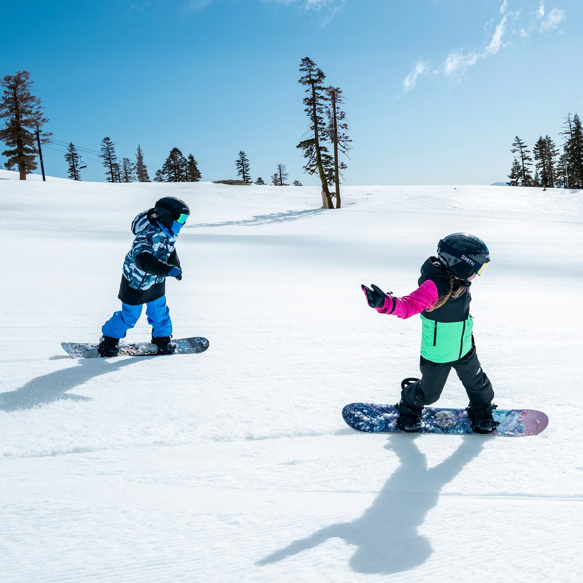 Two young snowboarders riding a groomed run, one in a blue and white patterned jacket and the other in a pink and green jacket, with shadows stretching across the snow and a chairlift line in the distance.