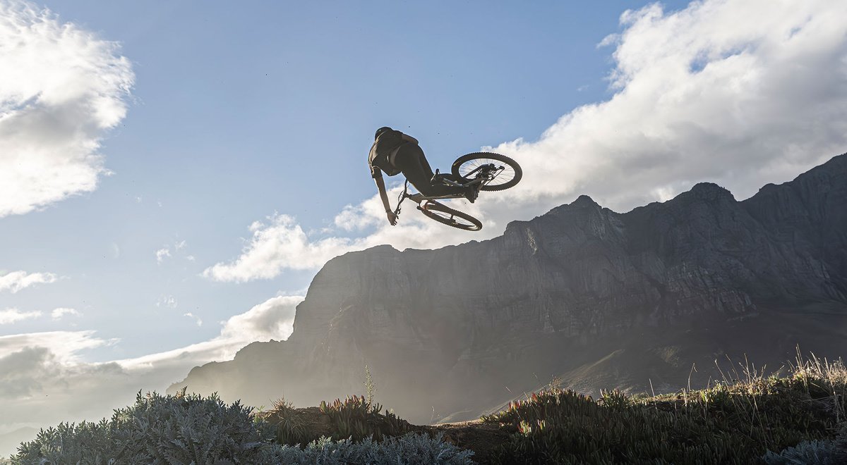A mountain biker performs an aerial trick, with dramatic mountain cliffs and clouds in the background.