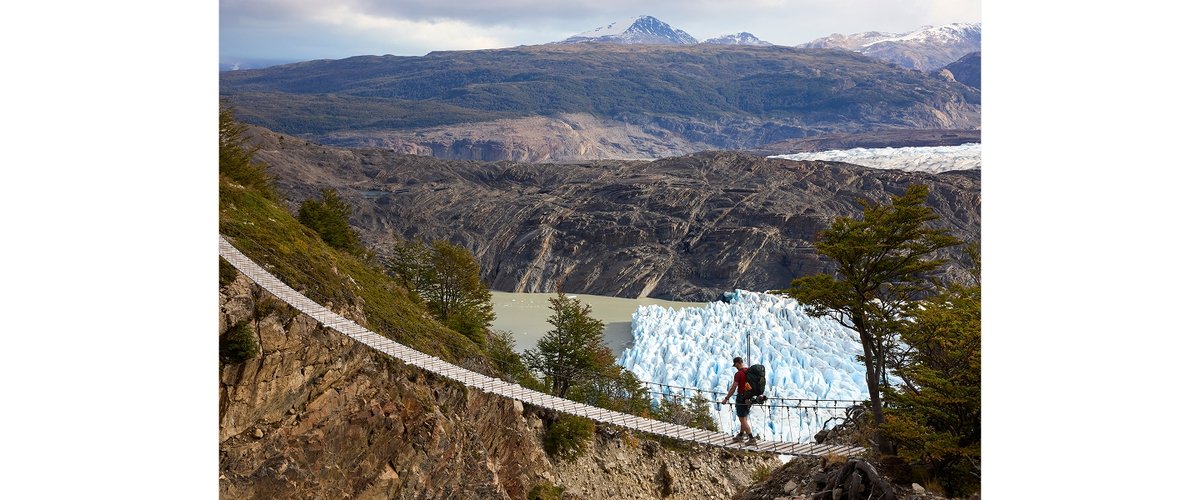 A hiker walks on a suspension bridge. Under them, a glacier can be seen.