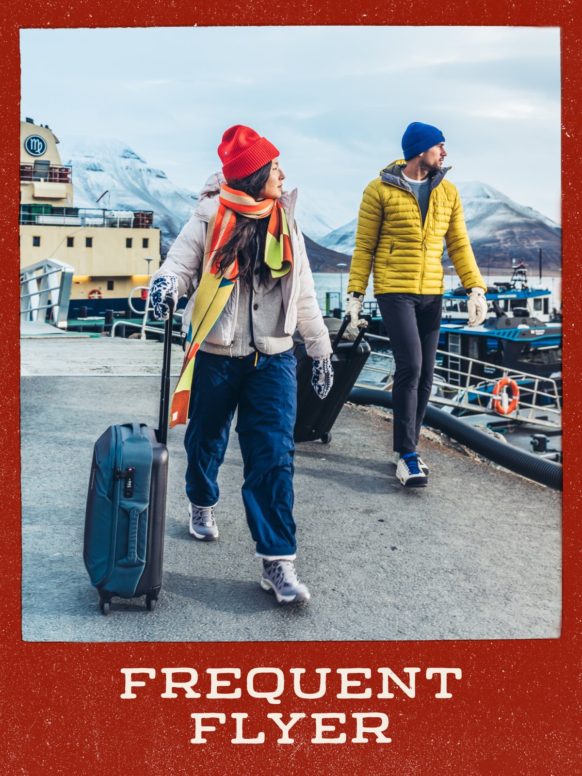 Two travelers pulling wheeled carry-on suitcases along a harbor dock with snowy mountains behind them: “Frequent Flyer.”