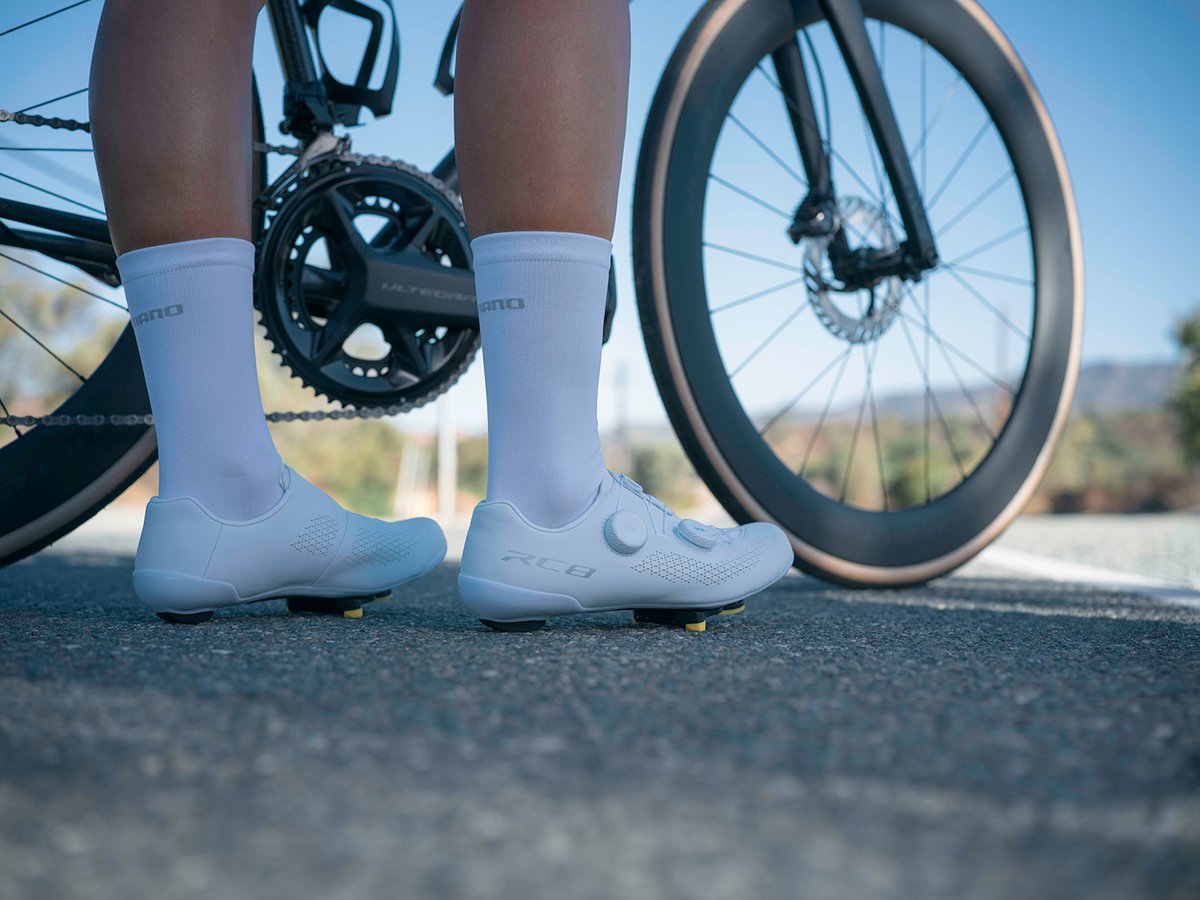 Cyclist's feet in white RC810 shoes standing beside a bike, Ultegra crank blurred in the background, road setting