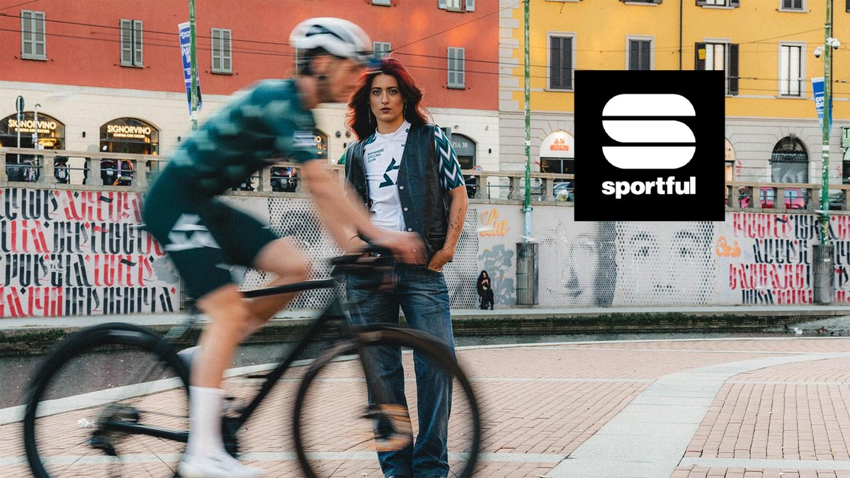 Urban cyclist in motion riding past a woman standing in a colorful European town square wearing sportswear and a cycling helmet.