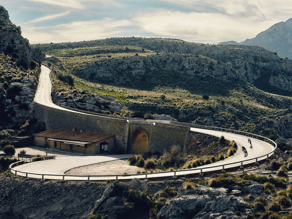 Three road cyclists descending a serpentine mountain road past a stone tunnel portal and building, with scrub-covered hillsides receding into the distance.