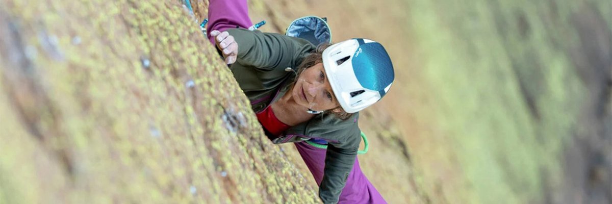 Rock climber wearing purple climbing pants and white helmet uses blue rope while ascending a natural stone cliff face.