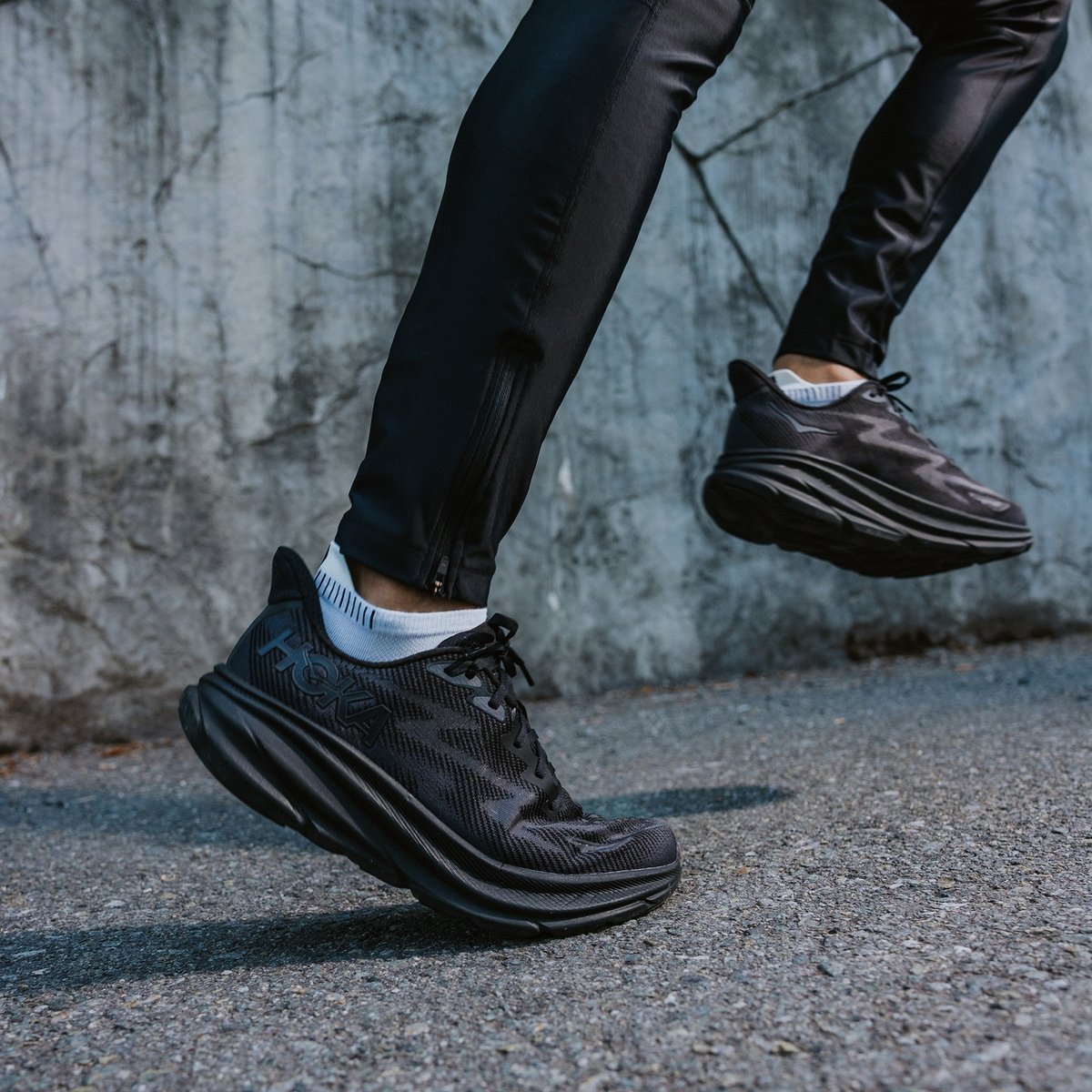 Closeup of a person in black HOKA shoes and black running tights running up a paved road.