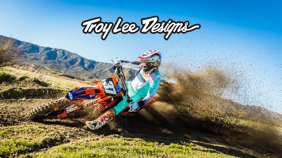 A motocross rider in colorful Troy Lee Designs gear performs a dramatic dirt bike maneuver on a hillside course with mountains in the background.
