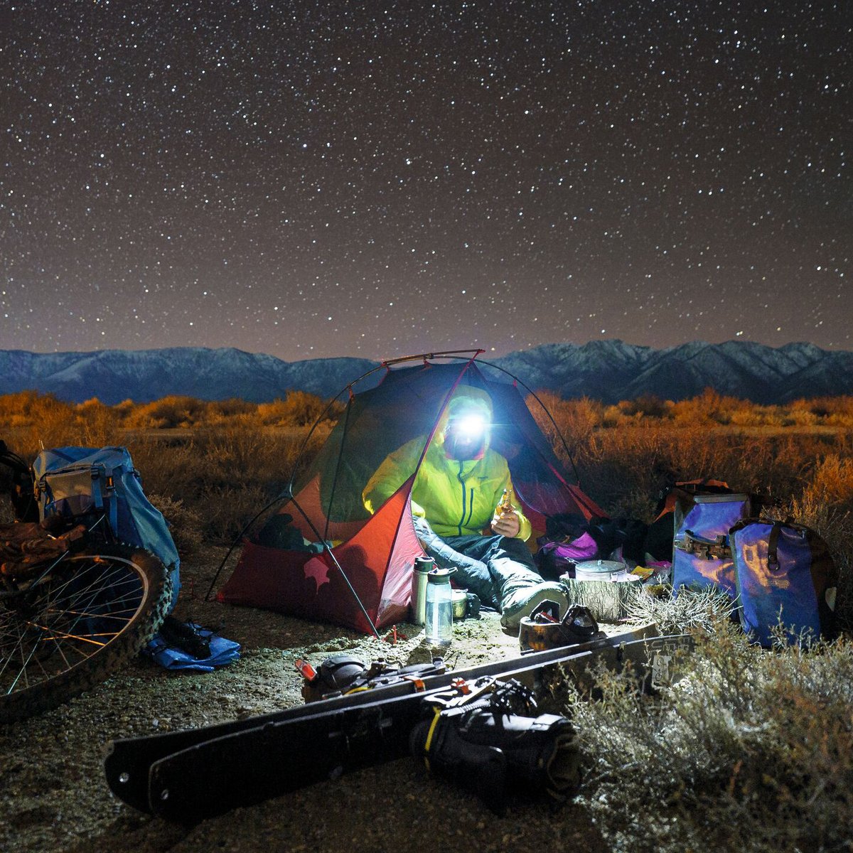 Camper enjoys a night under the stars in a brightly lit tent surrounded by outdoor gear, mountains, and desert landscape.