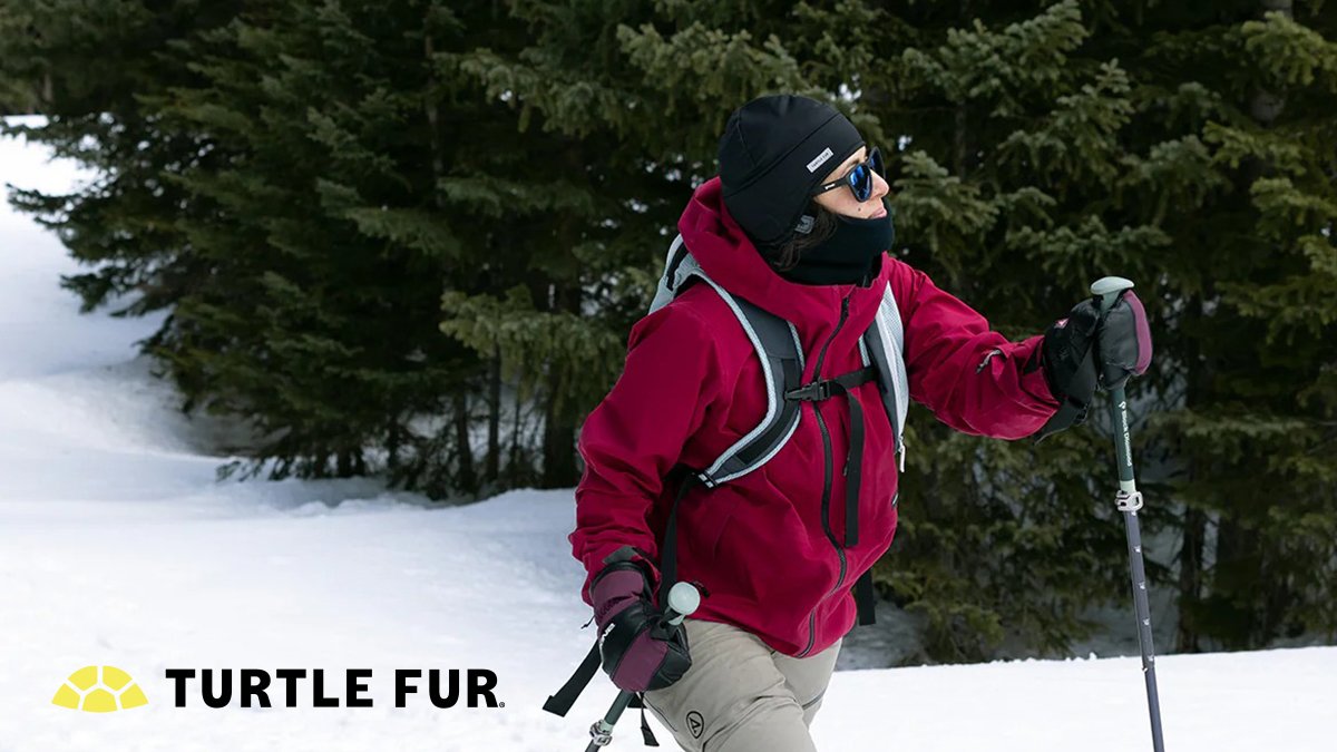 A hiker wearing a Turtle Fur beanie and neck gaiter treks through a snowy forest with trekking poles.