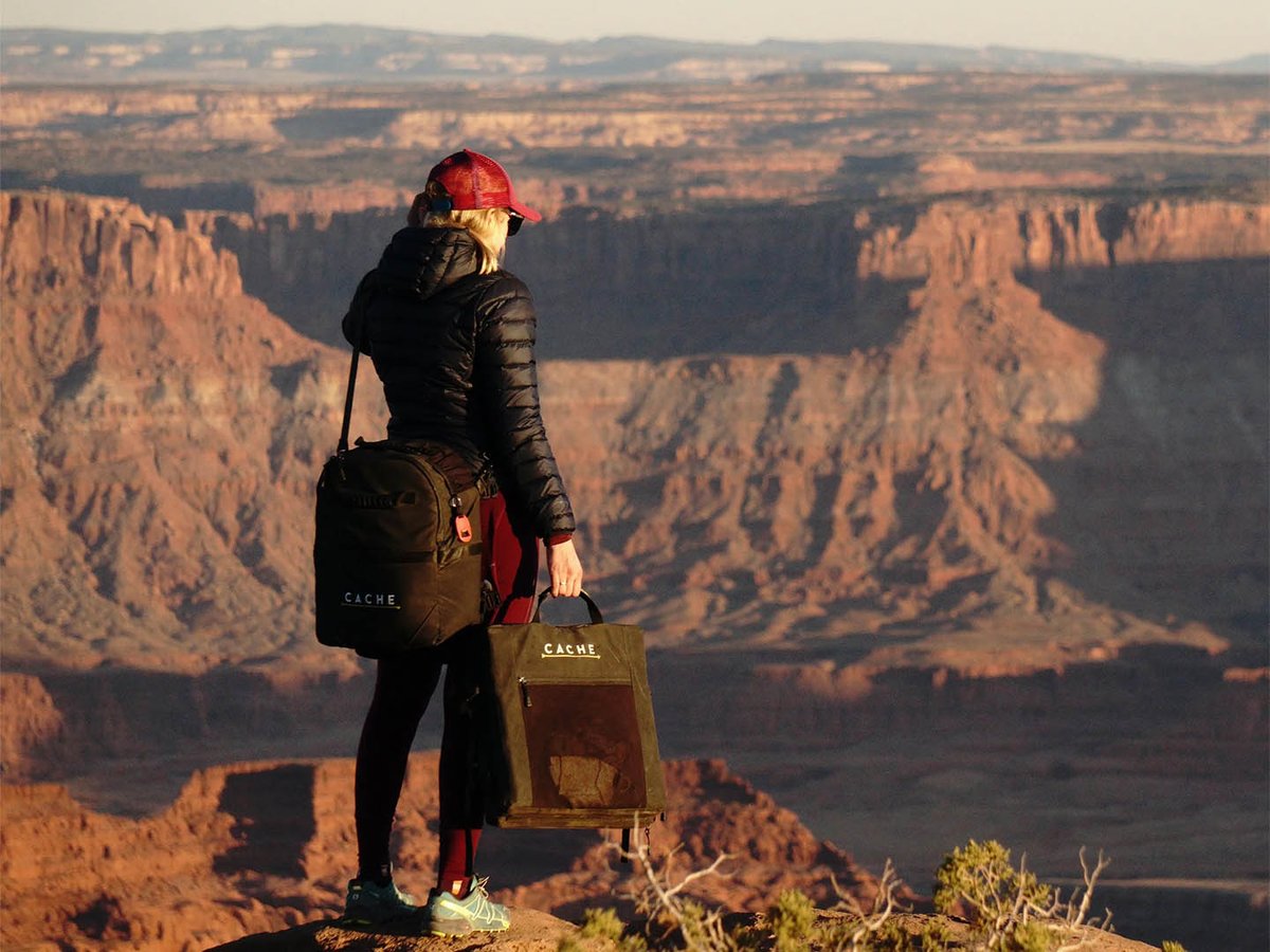 Woman at a canyon overlook carrying a CACHE backpack and matching CACHE tote, red rock mesas stretching to the horizon.