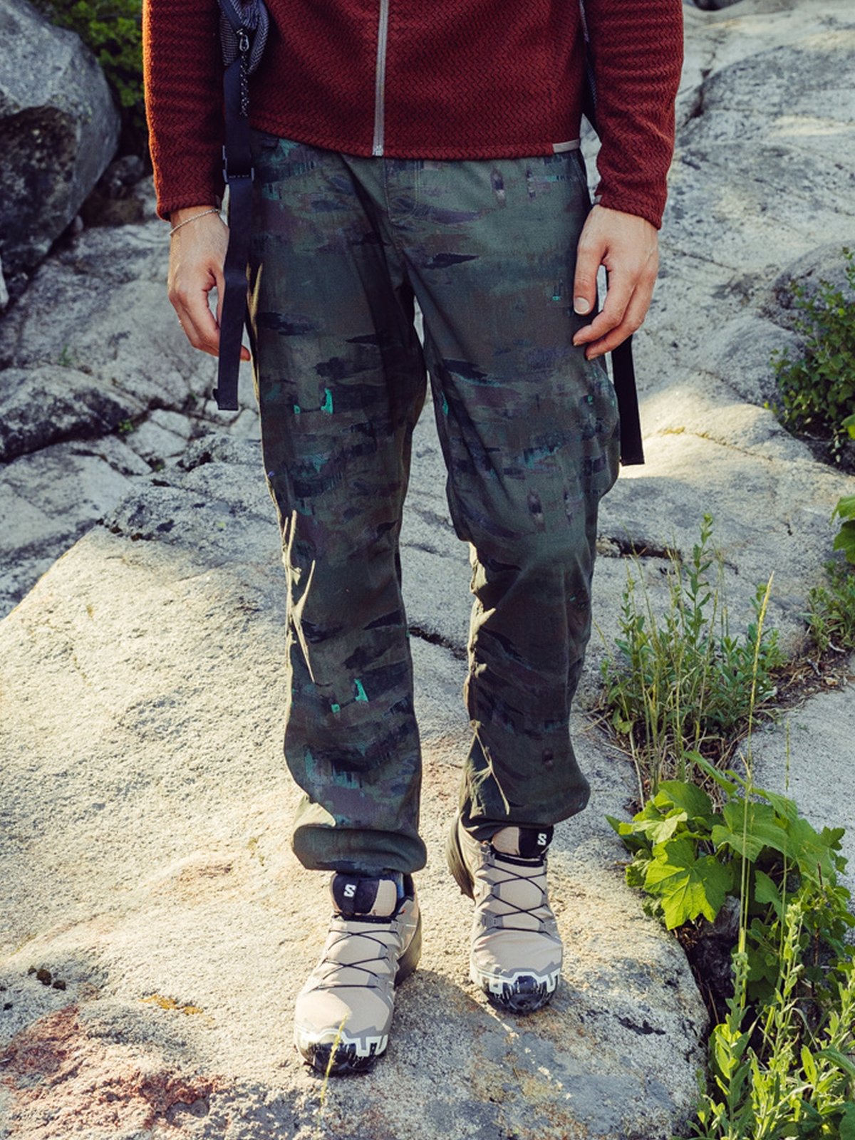 A person stands on a rocky outdoor surface wearing a textured red jacket, green camouflage cargo pants, and light-colored hiking shoes.