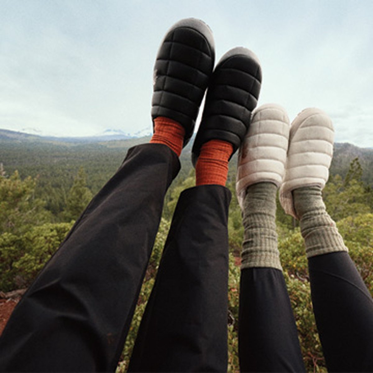 Two pairs of feet wearing black and white puffer slippers are propped up against a scenic forest background.
