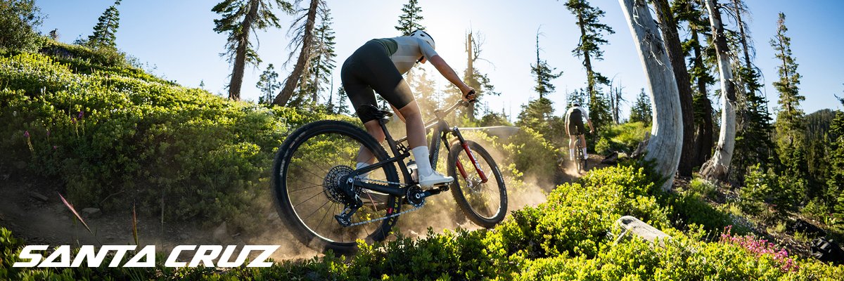 A rider carves a dusty singletrack turn on a Santa Cruz mountain bike through sun-drenched alpine shrubs, another rider visible ahead.