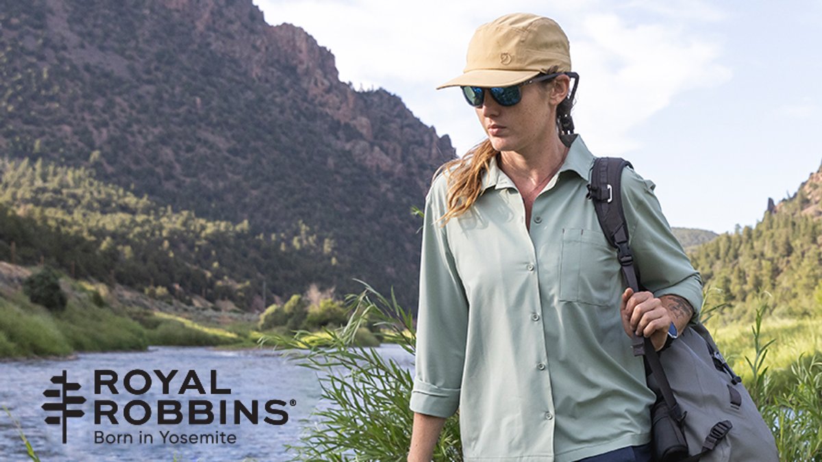 Woman wearing Royal Robbins outdoor clothing and gear standing by a mountain river in a scenic valley landscape.