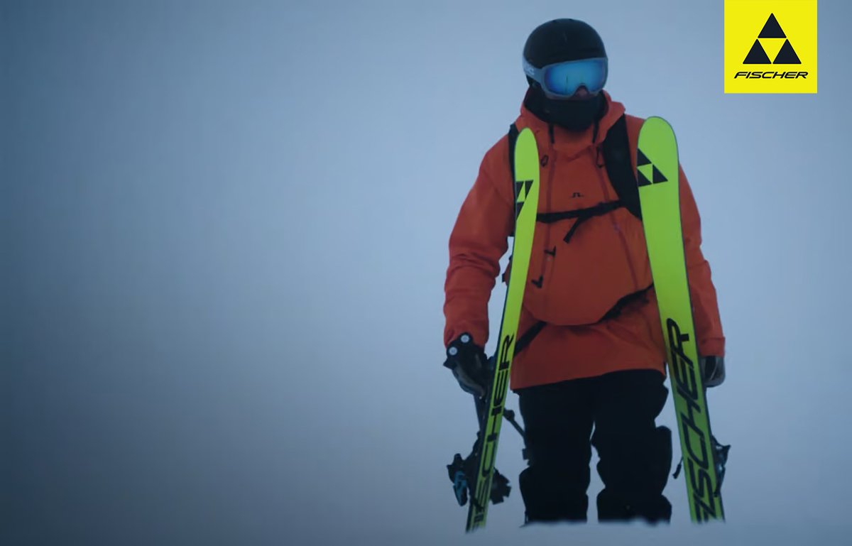 Alpine skier in red Fischer jacket carrying neon green Fischer skis against blue sky background.