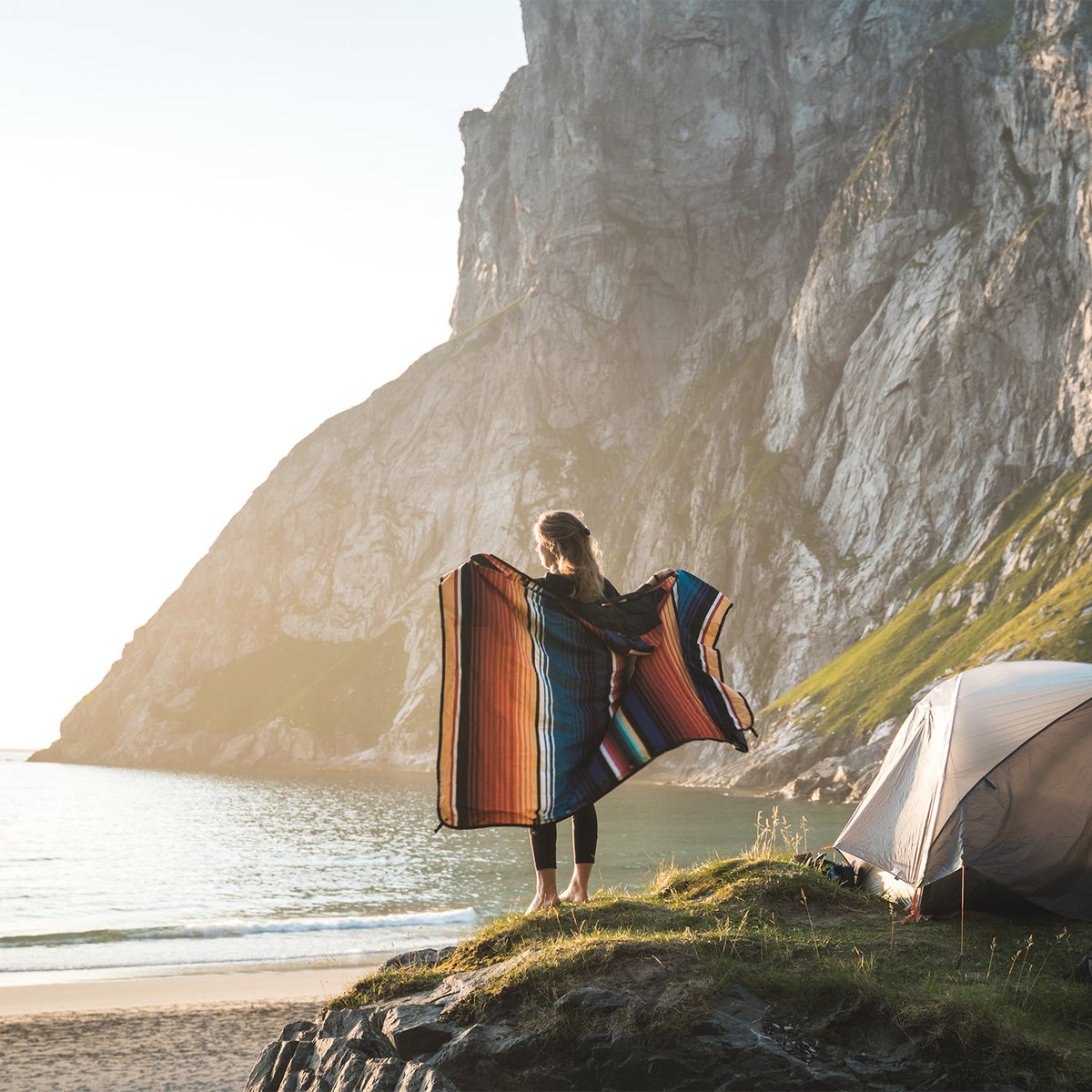 Peson standing holding up Coalatree Kachula blanket standing on rocky ocean overlook with a tent and rocky cliff in the background.