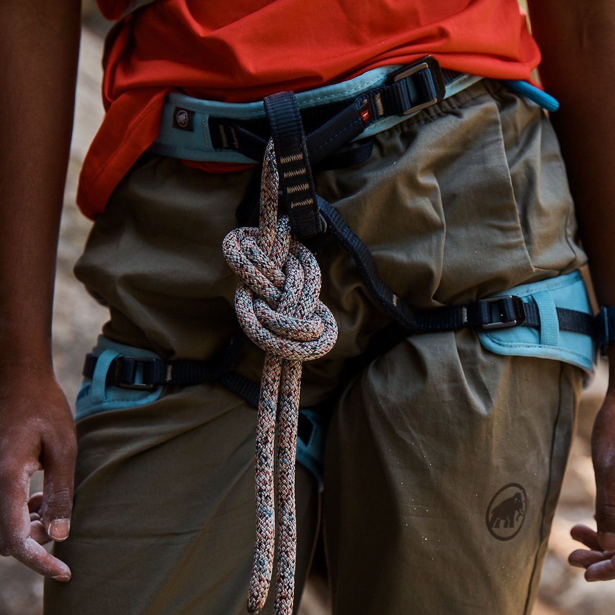 A close-up of a climber in an orange shirt with a blue harness and knotted rope.