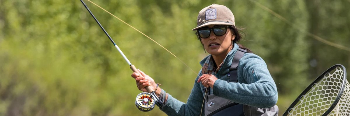 Fly fisherman wearing waders and holding a fishing rod with net in hand while standing in a river surrounded by forested landscape.