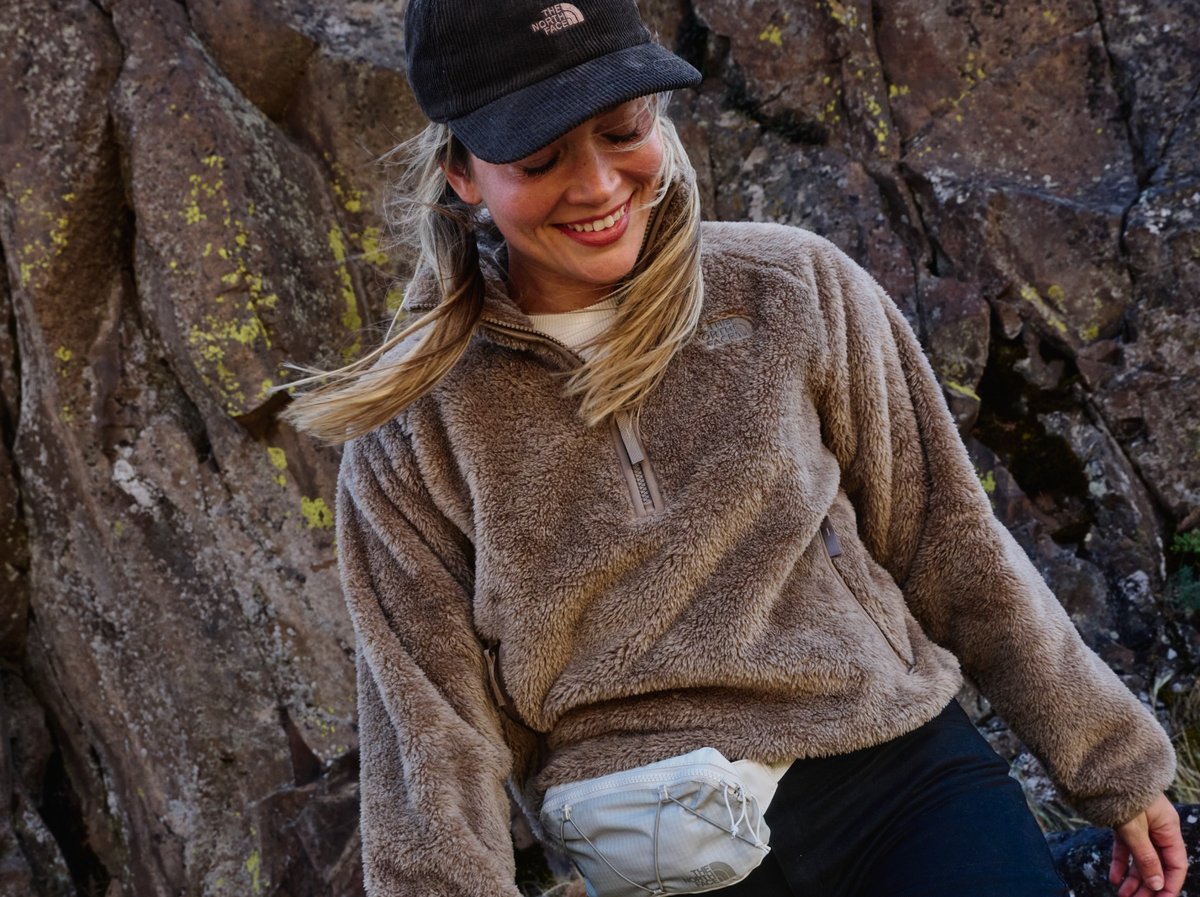 Smiling hiker in a North Face sherpa fleece and corduroy cap leans against lichen-covered granite, fanny pack at the waist.