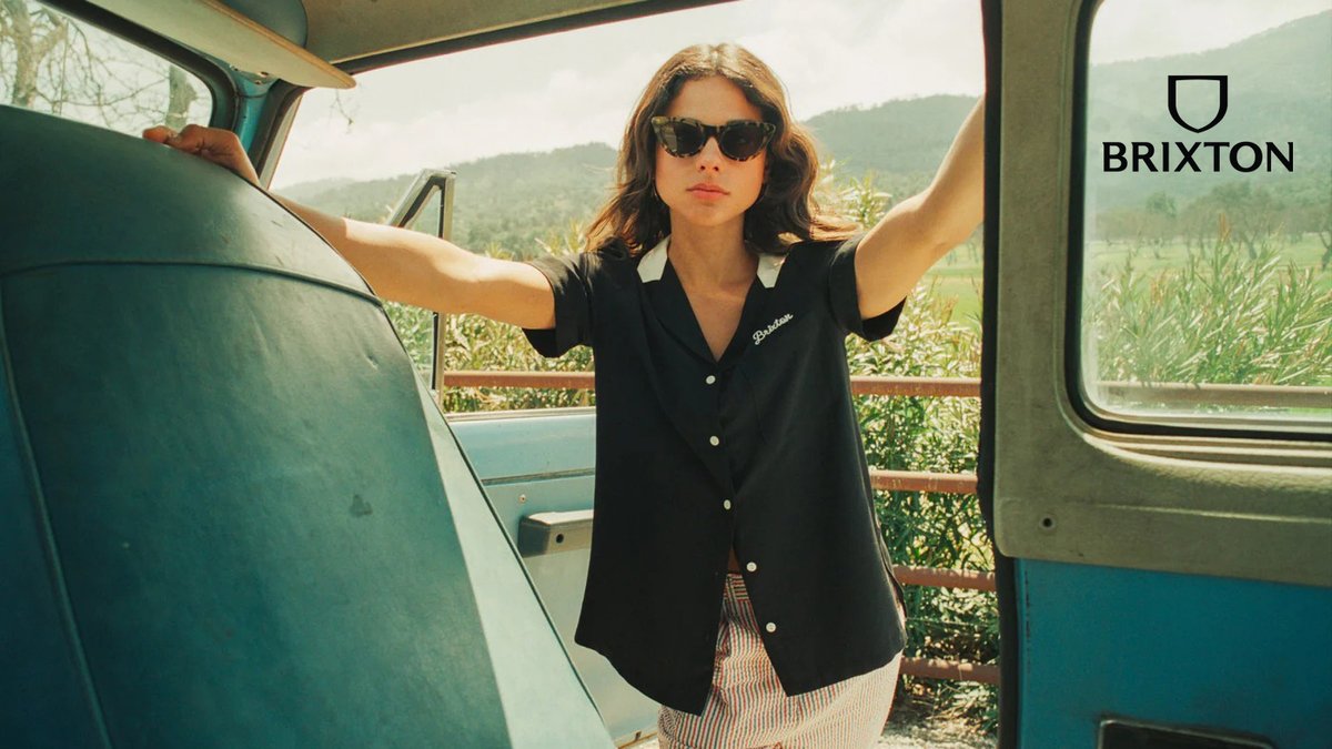 Woman wearing Brixton sunglasses and black blazer standing in vintage van with scenic mountain landscape visible through windows.