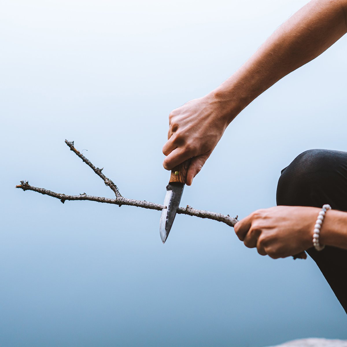 Person whittling a stick using Coalatree Haswell Knife with water in the background.