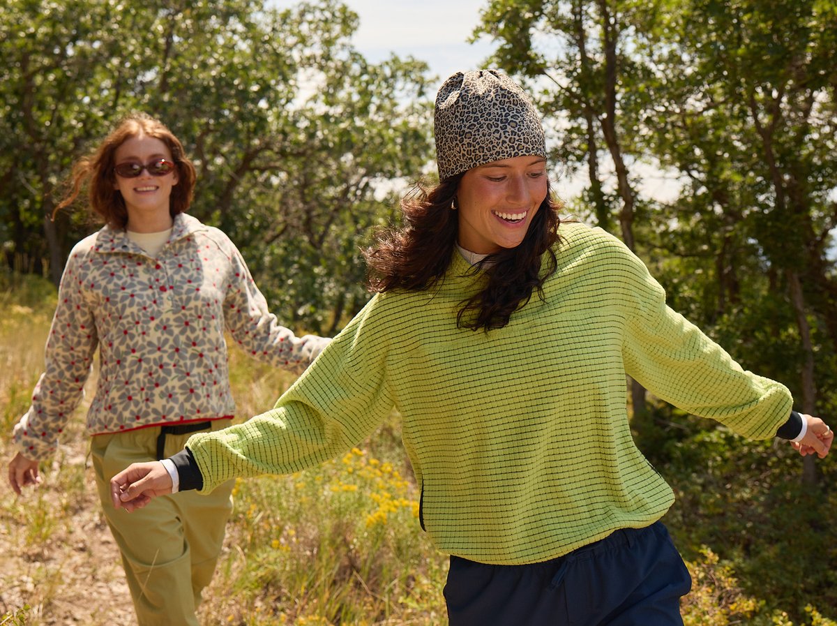 Two friends wearing patterned fleeces laugh and run down a sunny dirt trail through scrubby oak trees.