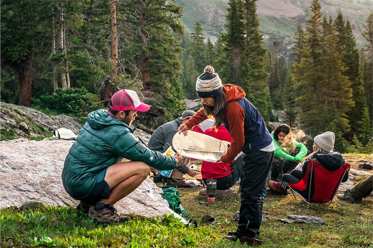Group of campers in a forested mountain setting refill water using the Platypus GravityWorks 4.0L filter system while others relax nearby.
