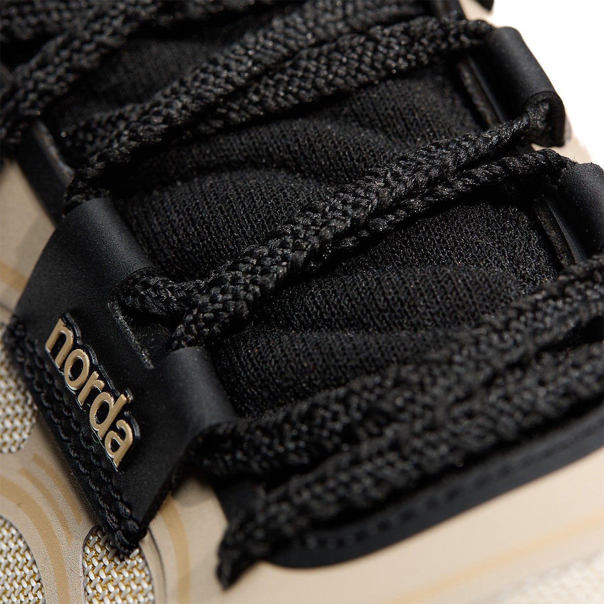 Close-up of the Norda lace closure system showing black laces threaded through a black branded lace lock with gold "norda" text, over a woven Dyneema upper.