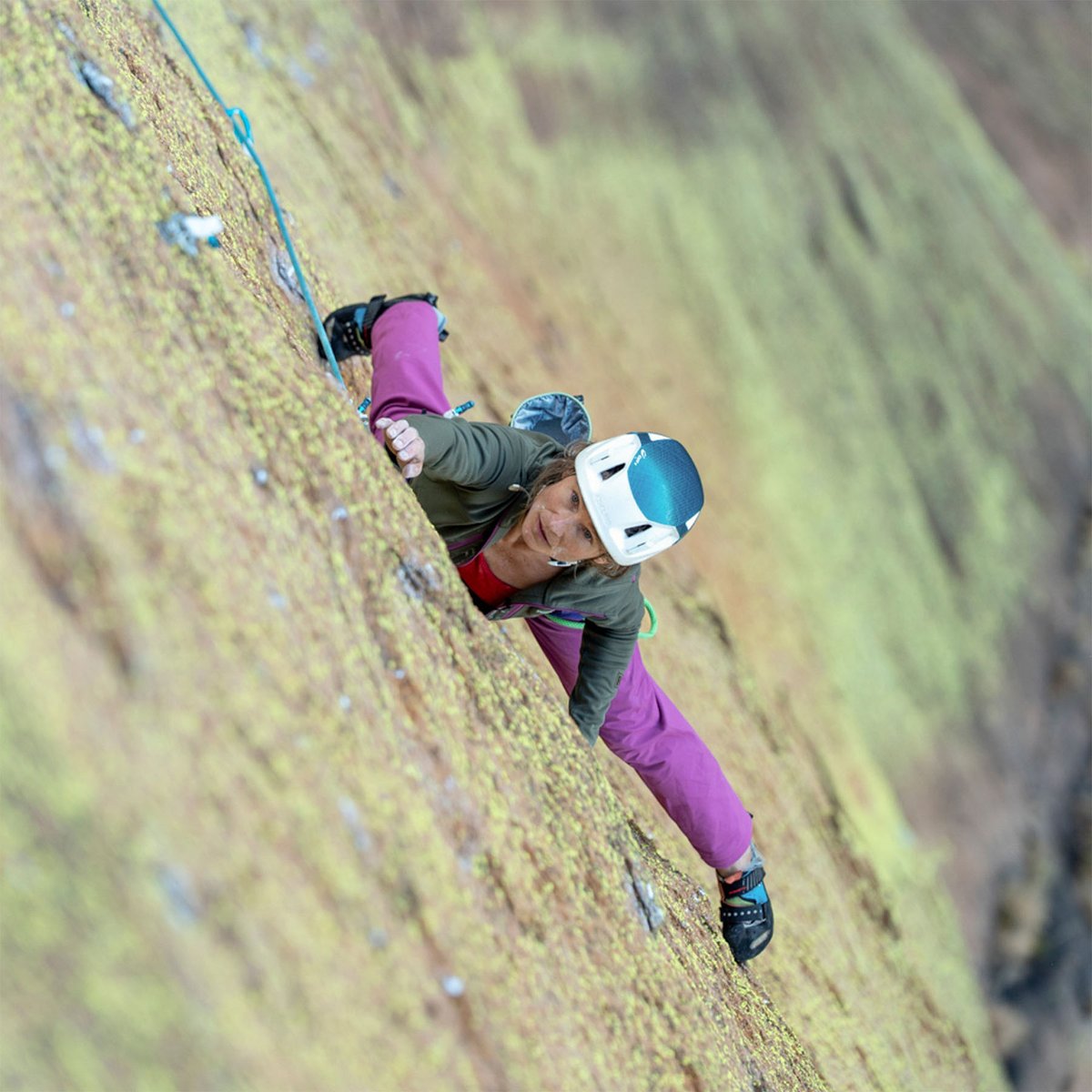 Climber wearing helmet and climbing shoes on lichen-covered granite slab with rope protection.