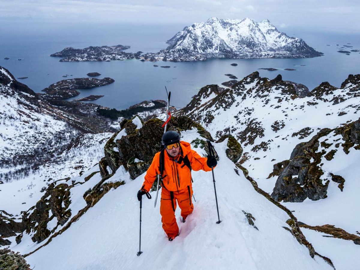 A skier in bright orange winter gear ascending a snowy mountain ridge with snow-covered peaks and blue arctic waters dotted with islands in the background.