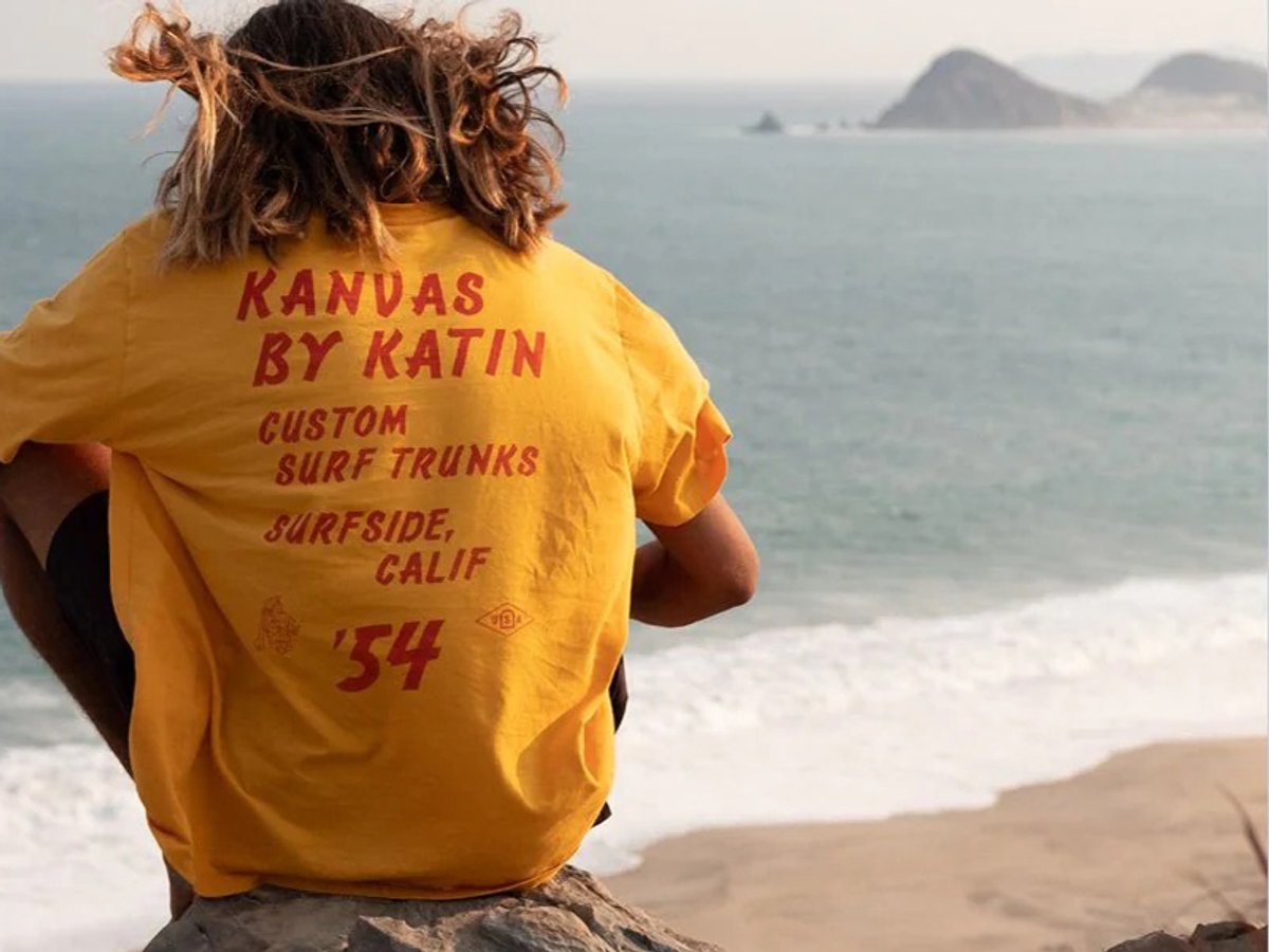 A person sits on a cliff overlooking the ocean wearing a yellow t-shirt with "Kanvas by Katin" printed on the back.