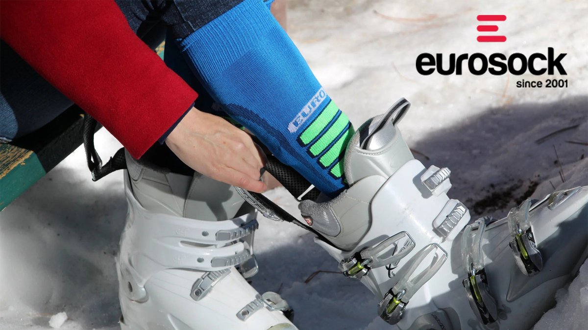 A person sitting on a bench adjusts their white ski boot to show a blue and green Eurosock.