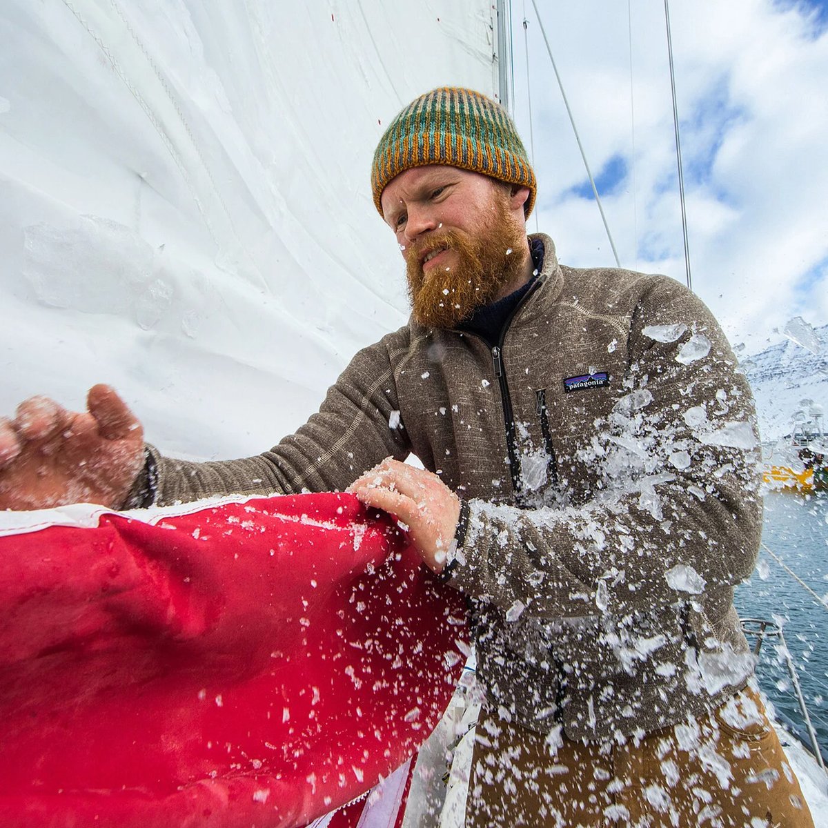Bearded man wearing a mustard beanie and Patagonia fleece jacket holds a red sail covered in snow while working on a sailboat in cold weather conditions.
