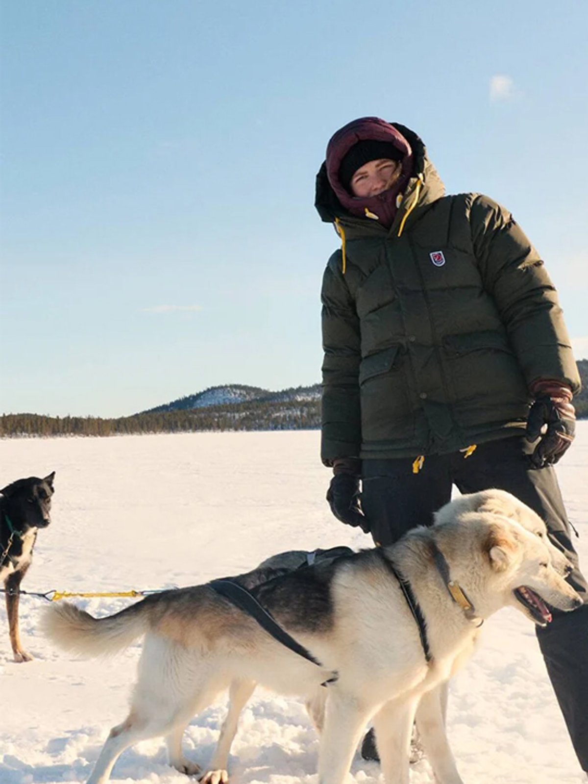 A woman wears a Fjallraven jacket with the hood up. Sled dogs stand at her feet.