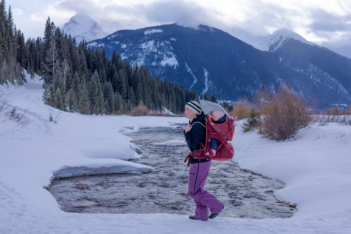 A woman walks near a snowy river carrying a child in a red Osprey carrier.