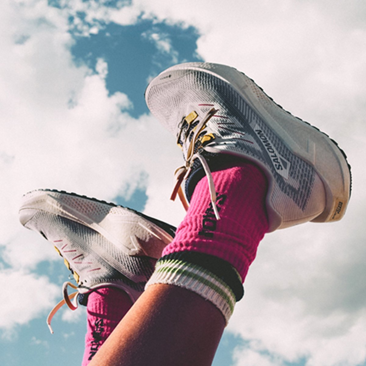 A pair of legs wearing white Salomon running shoes and bright pink socks with green and white stripes held up in the air, seen before the scattered clouds and blue sky as if their person is laying on their back. 