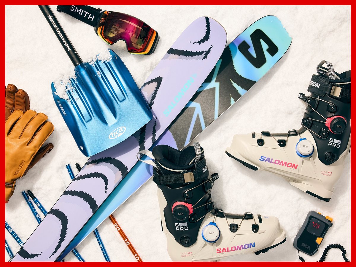 Ski gear displayed on snow, including Salomon skis and boots, Smith goggles, Hestra gloves, and a BCA shovel, probe, and beacon.