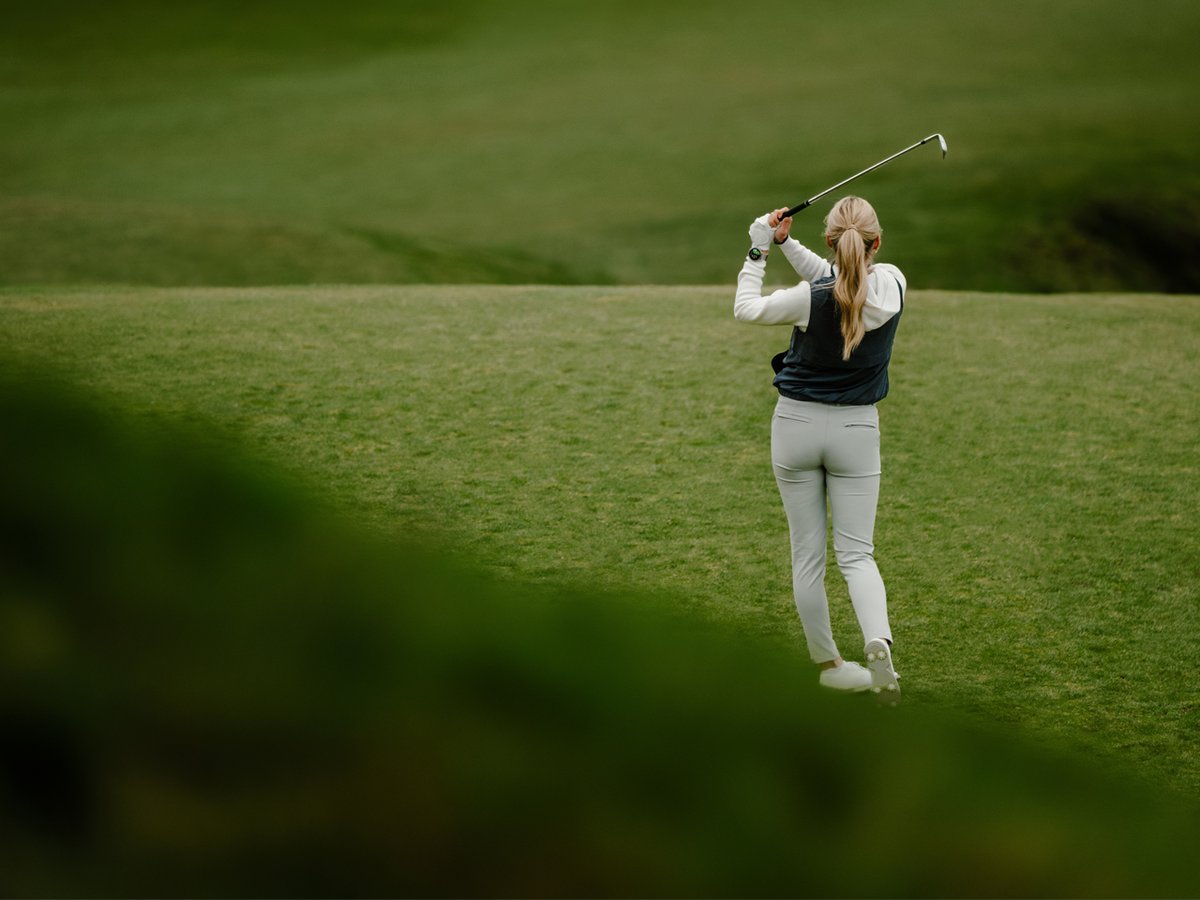 A woman faces away, mid-swing of her golf club.