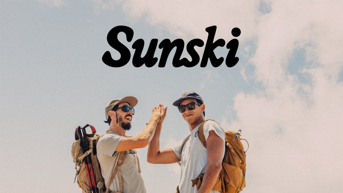 Two male hikers wearing sunglasses and backpacks high-five against a clear blue sky while on a mountain adventure.