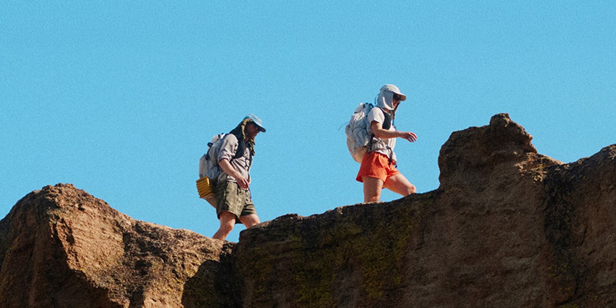Two hikers traversing a sun-exposed sandstone ridgeline with loaded packs against a clear blue sky.