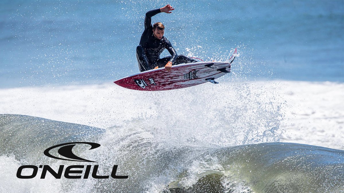 A surfer in a black wetsuit and sunglasses rides an air on a red and white surfboard above a powerful wave under clear blue skies.